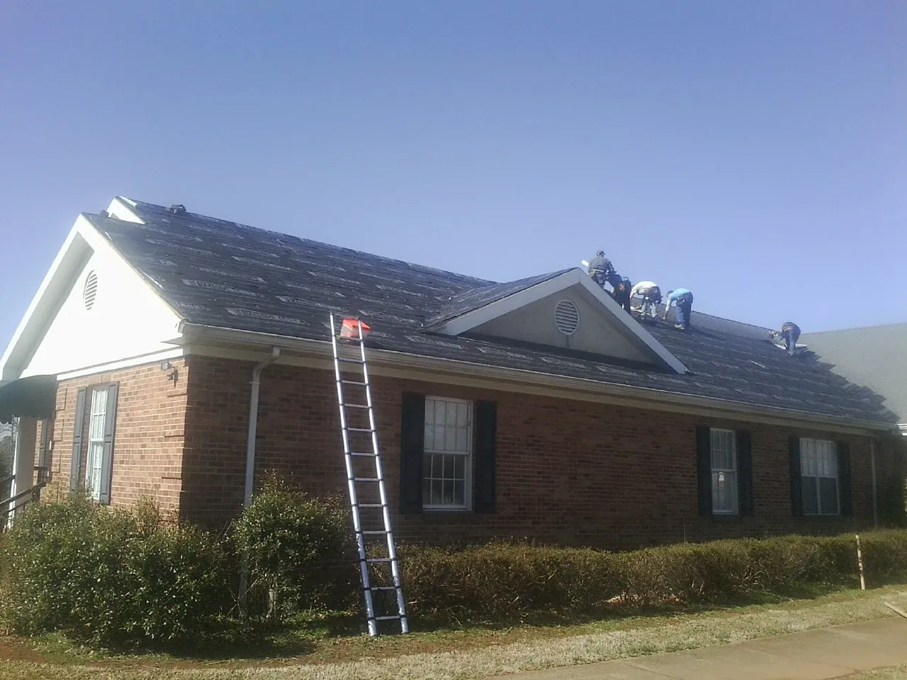 Crew tearing shingles off a brick ranch roof with a ladder set — Metal Roof Repair work by our crew in Rock City Falls