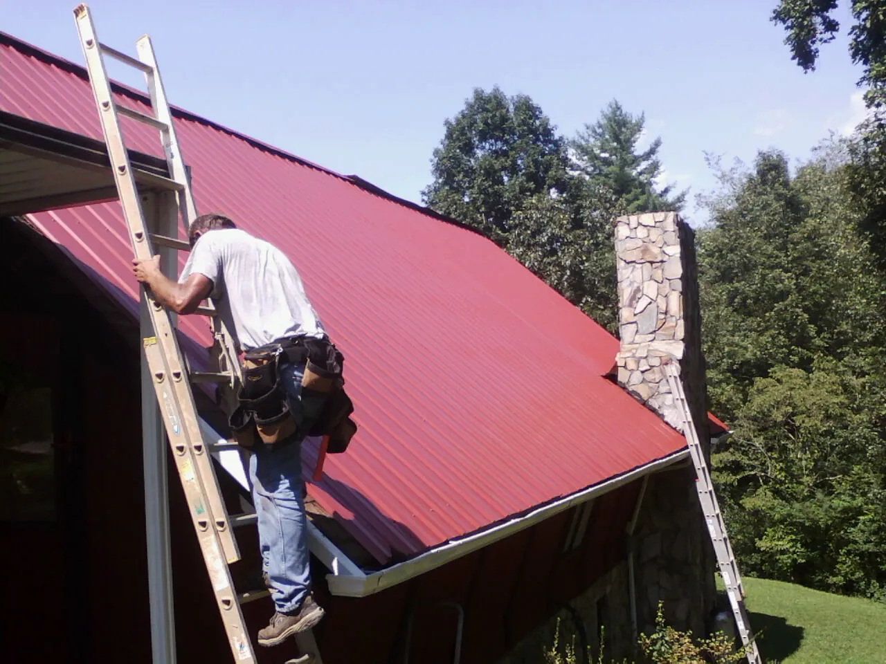 Roofer on a ladder beside a red metal roof and stone chimney — Soffit & Fascia Repair by Roofing Brothers in Rock City Falls
